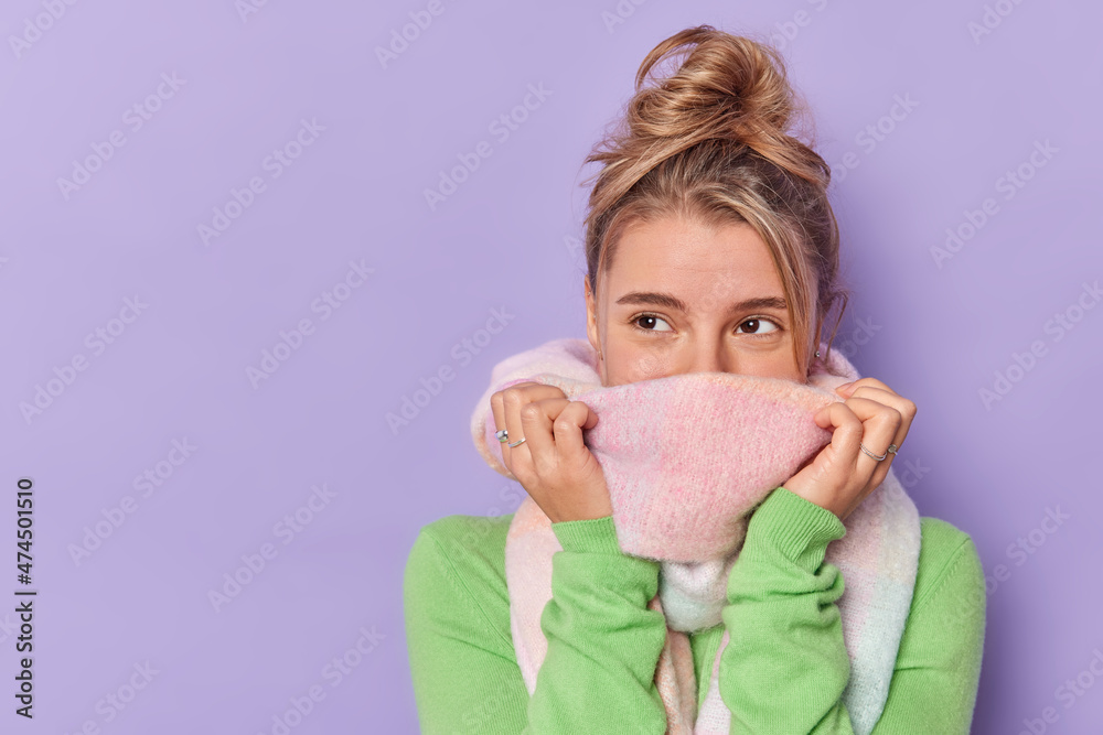 Thoughtful female model feels cold mouth with scarf tries to warm looks away poses against purple background blank copy space for your promotional content. People and winter clothes concept. Thoughtful female model feels cold mouth with scarf tries to warm looks away poses against purple background blank copy space for your promotional content. People and winter clothes concept.