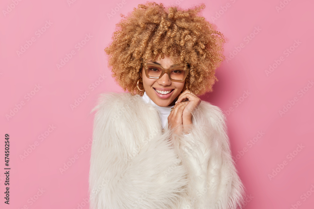 Portrait of cheerful lovely woman smiles happily keeps hands near face looks directly at camera wears eyeglasses and white coat feels good isolated over pink background. Fashion and people concept Portrait of cheerful lovely woman smiles happily keeps hands near face looks directly at camera wears eyeglasses and white coat feels good isolated over pink background. Fashion and people concept