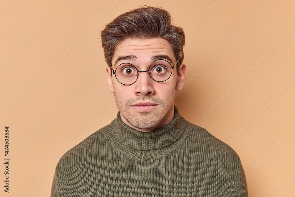 Portrait of shocked brunet European man stares surprisingly at camera keeps eyes widely opened wears round spectacles and sweater sees something breathtaking isolated over beige studio background Portrait of shocked brunet European man stares surprisingly at camera keeps eyes widely opened wears round spectacles and sweater sees something breathtaking isolated over beige studio background