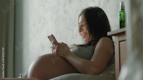 Young pregnant woman uses smartphone while lying in bed and laughing. Expectant mother is having fun using a mobile phone, watching funny videos. internet communication humor concept