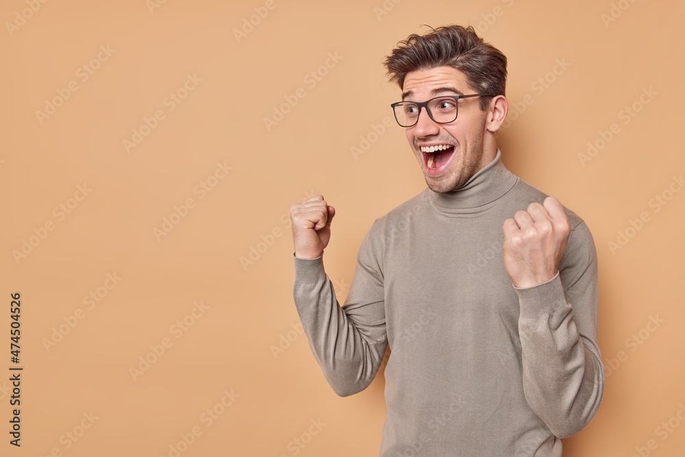 Overjoyed positive man clenches fists with triumph rejoices excellent news exclaims from happiness cannot believe in success wears casual jumper and spectacles gesture actively poses indoor.