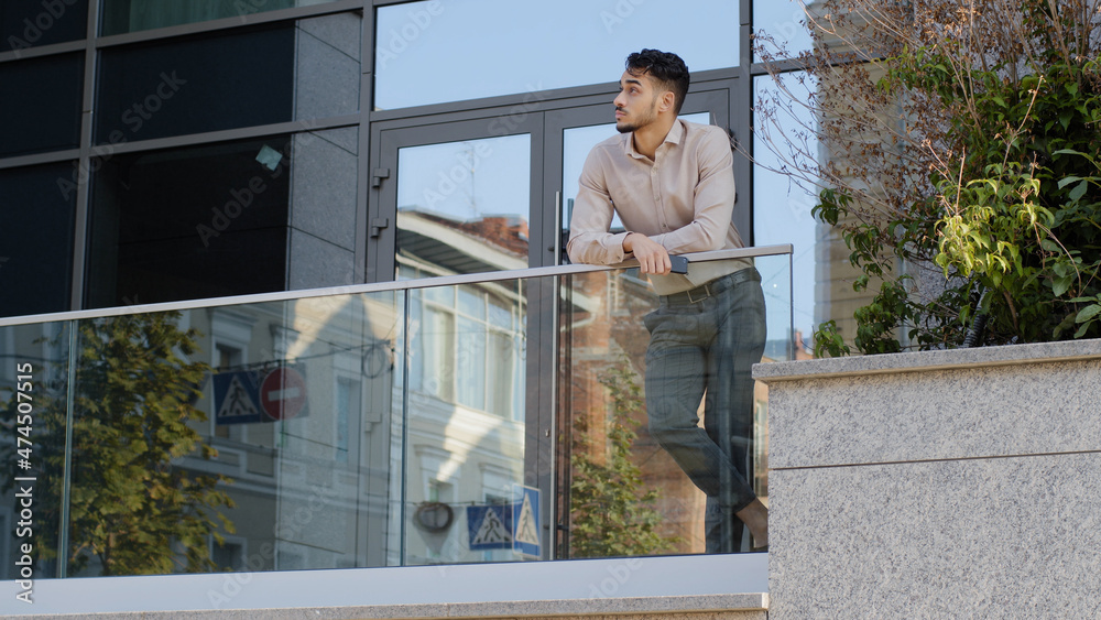 Confident young hispanic business man standing at railing modern skyscraper office building looking away holding phone thinking resting. Successful leader guy male on balcony terrace relaxing think