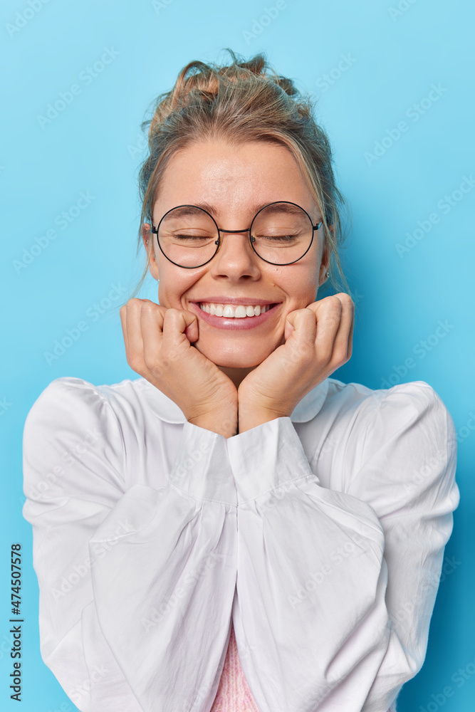 Beautiful young European woman keeps hands under chin smile toothily closes eyes wears round transparent glasses and white elegant shirt isolated over blue background. Happy emotions concept