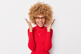 Waist up shot of emotional curly woman raises palms and screams from joy feels excited wears spectacles and red turtleneck isolated over white background. People emotions and reactions concept