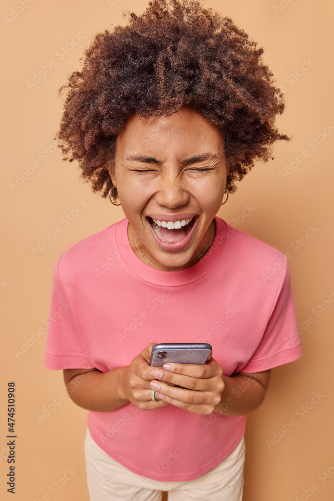 Indoor shot of emotional curly haired young woman uses mobile phone exclaims joyfully tests new smartphone application posts internet blog wears casual t shirt isolated over beige background
