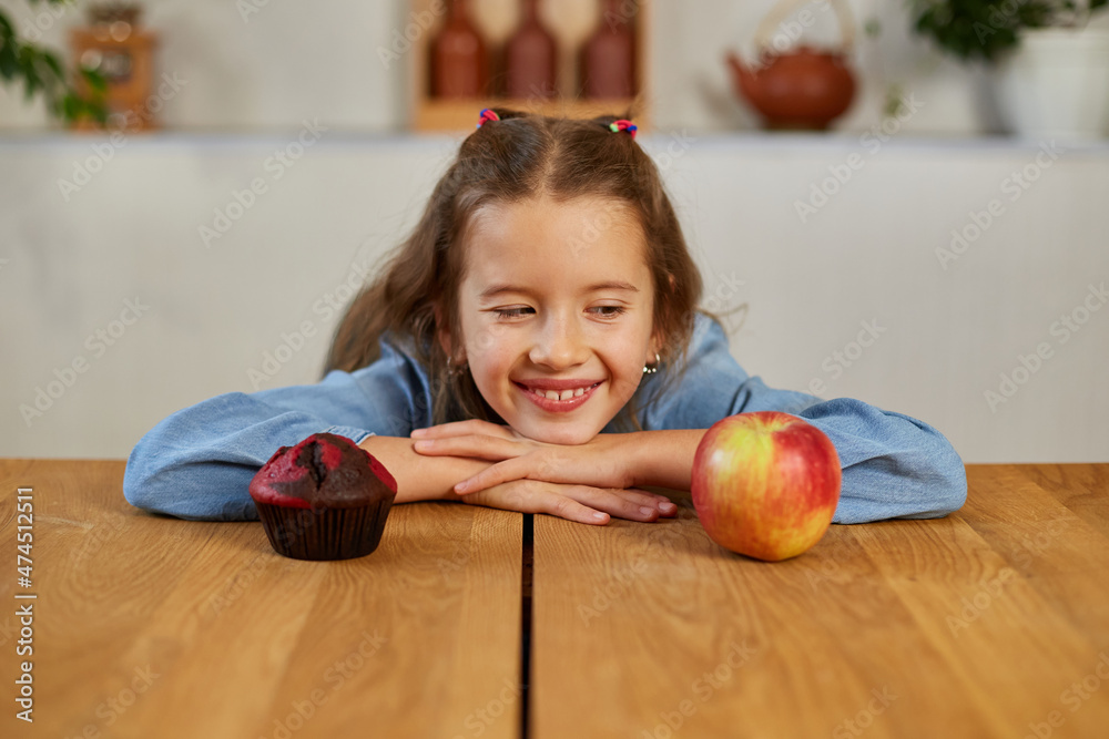 Fototapeta premium Little girl looking on sweet cake and fresh red apple and choosing in kitchen at home