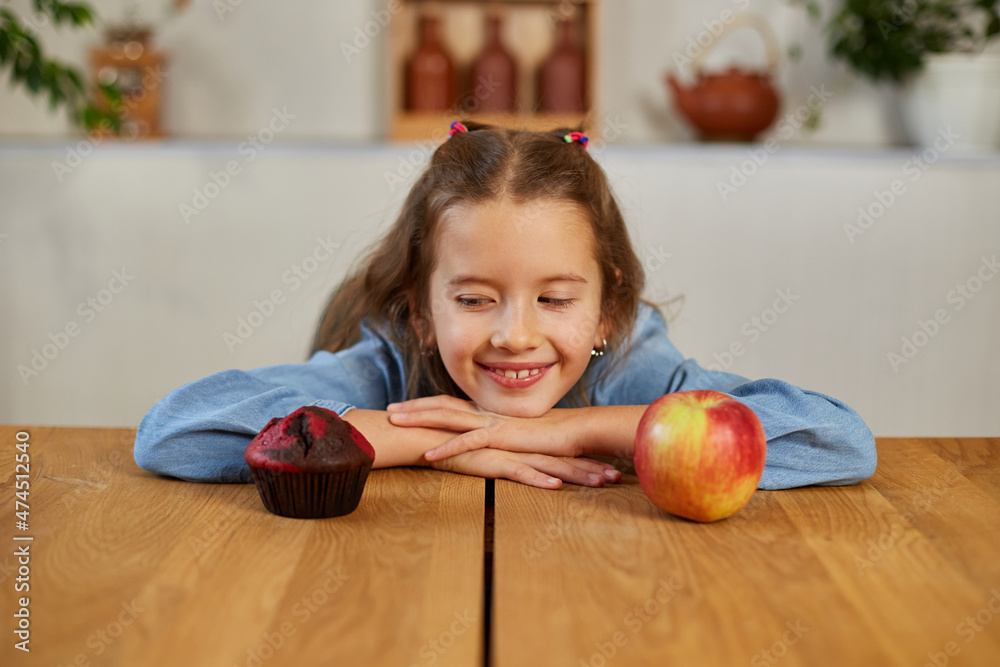 Fototapeta premium Little girl looking on sweet cake and fresh red apple and choosing in kitchen at home