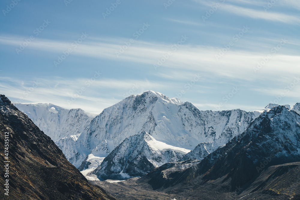 Alpine landscape with high snowy mountain with peaked top under cirrus clouds in sky. Atmospheric view to big snow covered mountains in sunshine. Black rocks and white-snow pointy peak in sunlight.