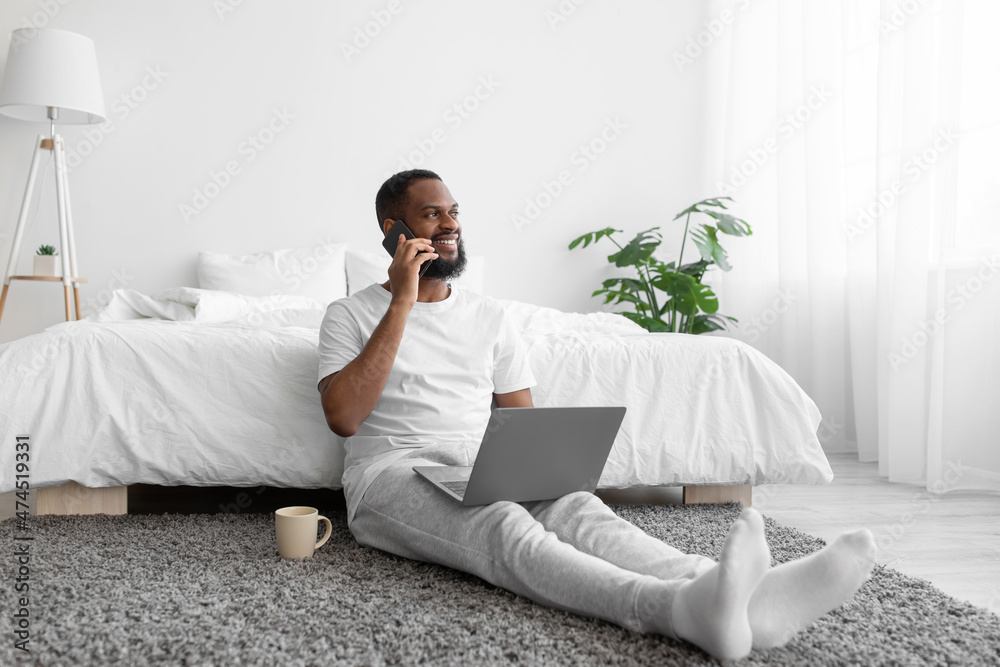 Glad young african american bearded guy sitting by bed working on laptop talking by phone with client Glad young african american bearded guy sitting by bed working on laptop talking by phone with client