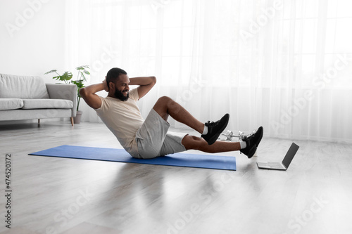 Happy young african american guy doing exercises for legs and abs on mat on floor in room interior