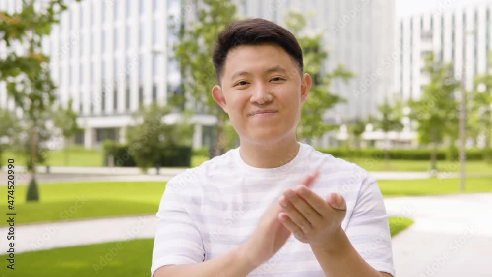 A young Asian man applauds to the camera with a smile in an urban area - a park and office buildings in the blurry background - closeup A young Asian man applauds to the camera with a smile in an urban area - a park and office buildings in the blurry background - closeup