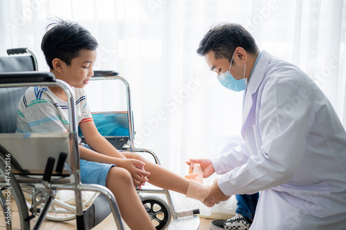 Asian boy sitting on a wheelchair is having the doctor wrap a cloth around his leg because he broke his leg at the hospital.