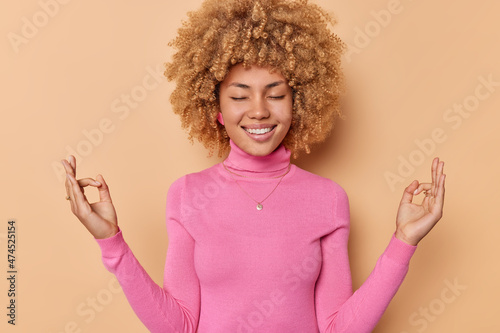 Horizontal shot of happy young woman with curly hair keeps eyes closed smiles broadly meditatees and makes zen gesture tries to reach nirvana smiles pleasantly wears pink jumper poses indoor