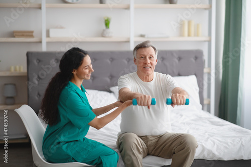 Young physiotherapist helping senior man to do physical exercises with dumbbells on bed at home