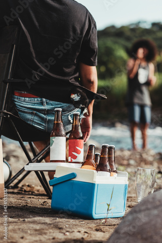 Vertical de una mujer desconocida sentada en una silla con  y una hielera con cervezas a la orilla de un río en Costa Rica y un hombre de fondo desenfocado