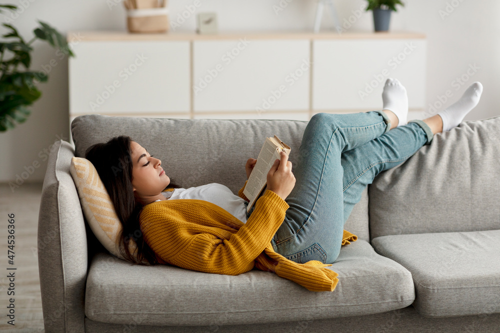 Cozy stay at home activities. Young arab woman reading her favorite book, lying on sofa in living room interior - obrazy, fototapety, plakaty 