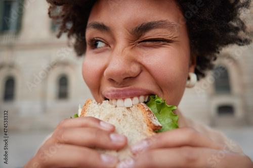 фотографія Close up shot of positive curly haired glad young woman winks eye holds appetizing sandwich with hands bites delicious snack returns from fast food restaurnat poses outdoors