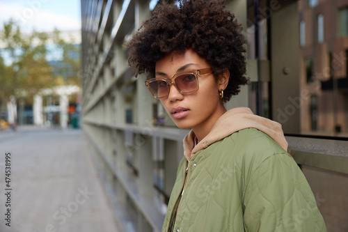 Horizontal shot of serious curly haired young woman wears sunglasses and jacket walks in city poses against blurred urban background thinks about something. Urban lifetyle and spare time concept photo