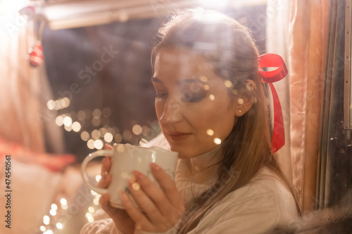 blurry portrait of candid woman face through the window glass holding in hands and enjoyment cup of cocoa drink and relaxing on happy new year and merry christmas eve. bokeh of glowing lights garland