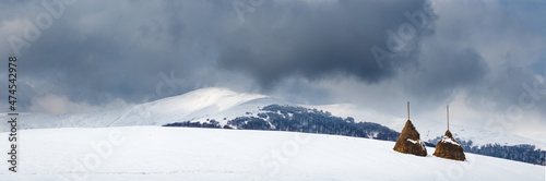 Wide panorama of snowy mountains under a dramatic cloudy sky. The rural area of Carpathian mountains with haystacks. © stone36