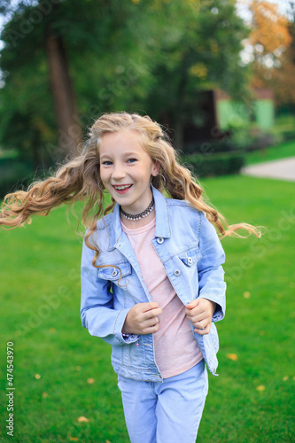 A positive blue-eyed, blond-haired girl in a denim jacket on a green lawn on a sunny day
