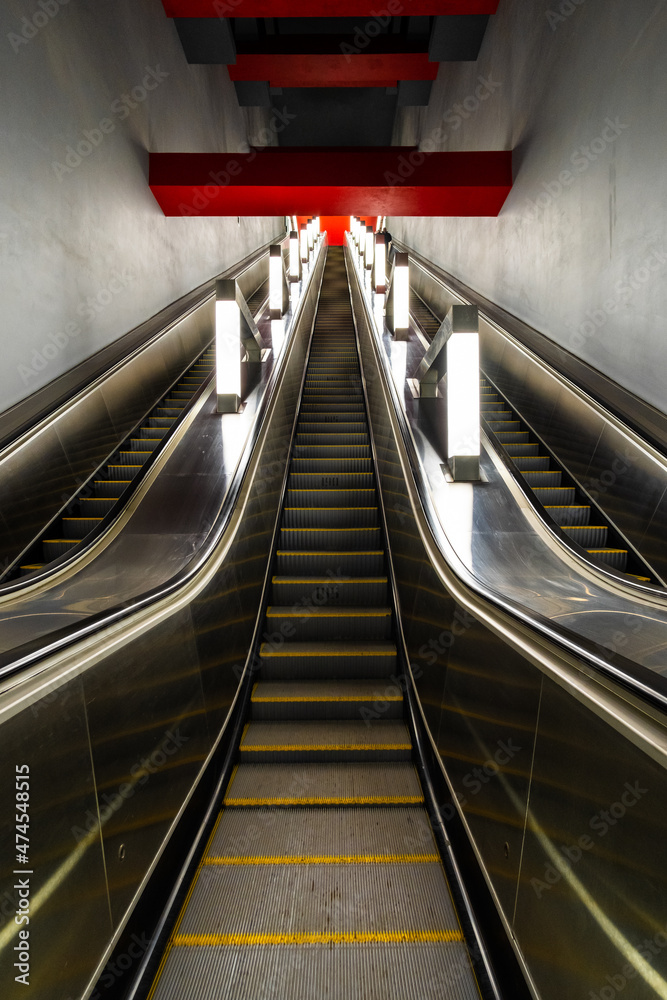Steps of a long escalator going up to the red slabs on the ceiling. Linear perspective. Vertical image.