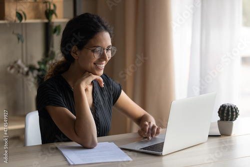Smiling young Latino woman in glasses sit at desk at home office work online on computer gadget. Happy millennial Hispanic female study distant on laptop, take course or training. Technology concept.