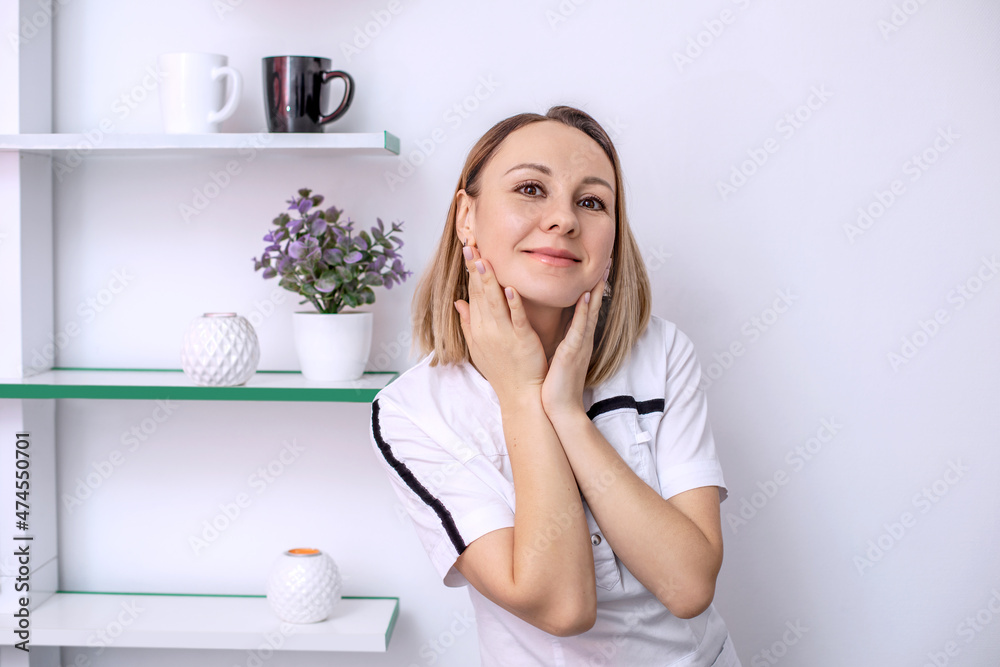A woman doctor or beauty master in a white coat in the office looks at the camera and smiles. Portrait of a young woman in light colors. Interior of a beauty salon or medical room.