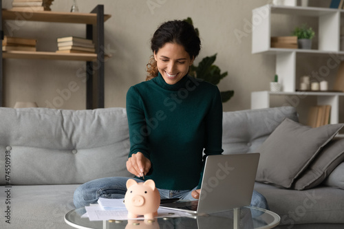 Smiling young Hispanic woman put coin into piggybank save money for future, pay bills taxes online on computer. Happy Latin female manage household budget finances, make investment in piggy bank.