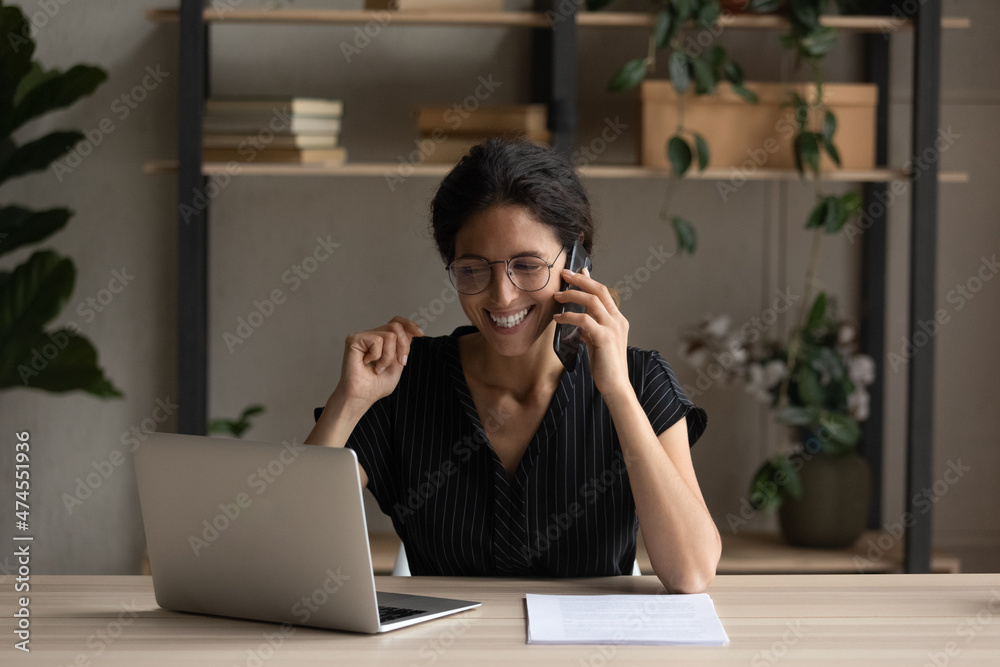 Smiling young Latin woman work online on computer at home office talk on cellphone with client. Happy Hispanic lady look at laptop screen consult customer on smartphone call. Freelance concept. Smiling young Latin woman work online on computer at home office talk on cellphone with client. Happy Hispanic lady look at laptop screen consult customer on smartphone call. Freelance concept.