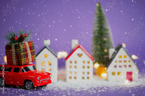 Christmas decor - red retro car on snow carries past houses with fairy lights in bokeh Christmas tree with gift boxes on roof. Toy on violet background. New Year greeting card. Cozy home © Ольга Симонова