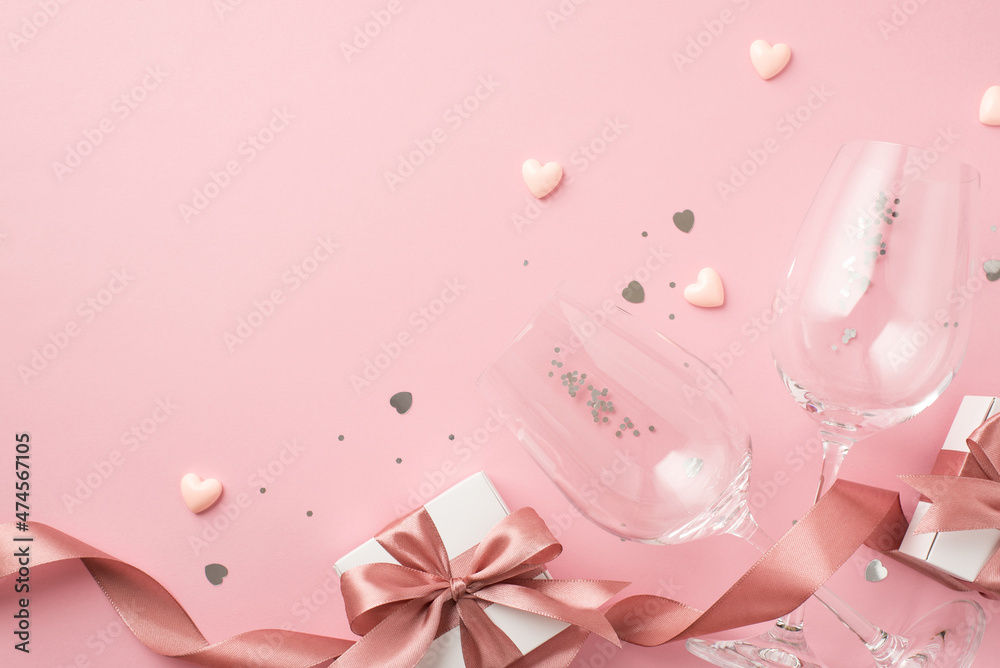 Top view photo of white gift boxes with pink satin ribbon bows small hearts two wineglasses silver sequins and heart shaped confetti on isolated pastel pink background with empty space