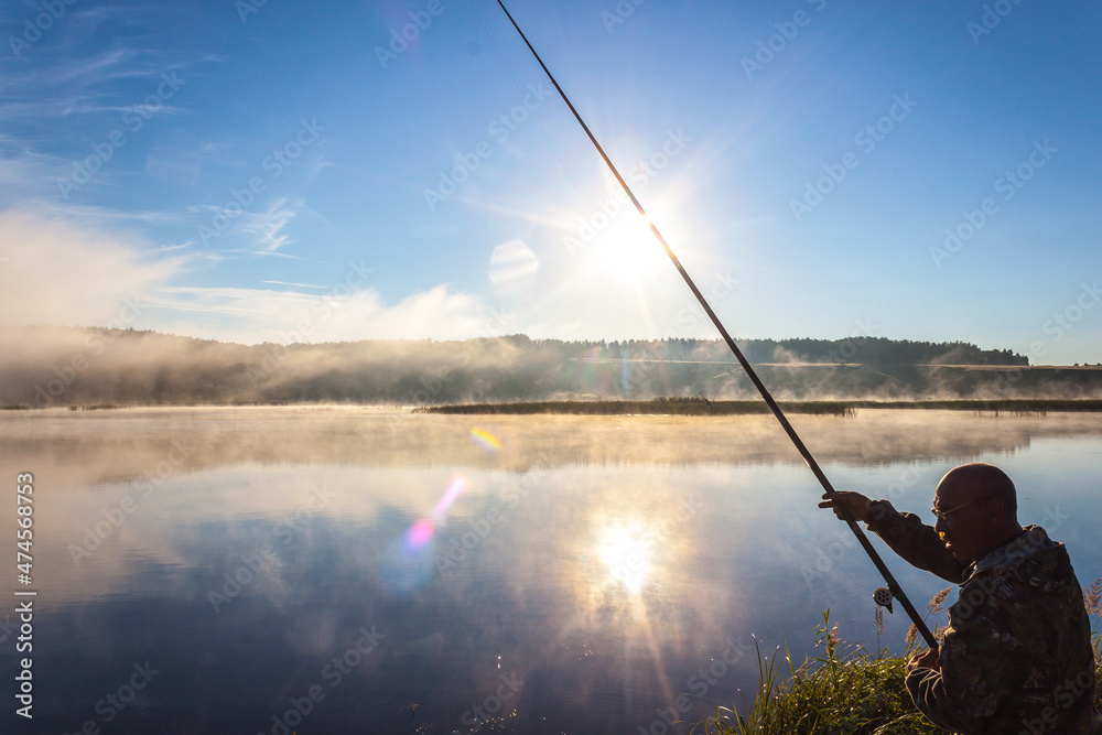 Fisherman fishing with spinning rod on a river bank at misty foggy sunrise