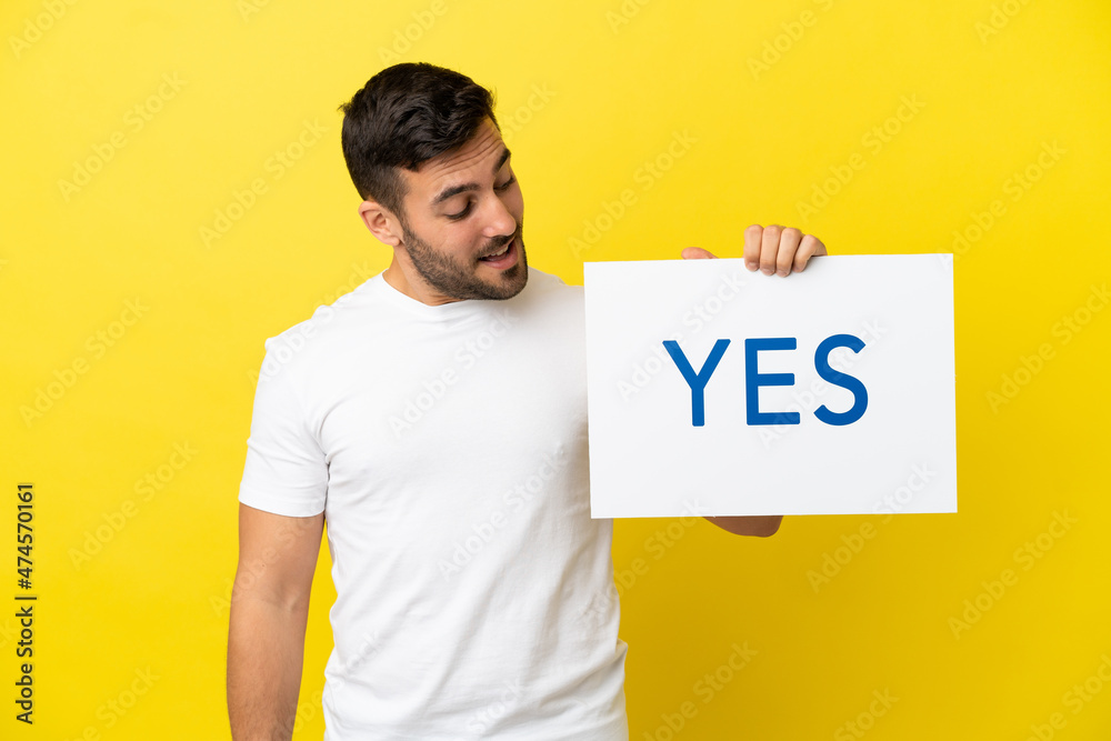 Young handsome caucasian man isolated on yellow background holding a placard with text YES Young handsome caucasian man isolated on yellow background holding a placard with text YES