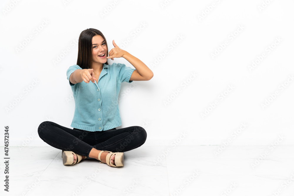 Teenager girl sitting on the floor making phone gesture and pointing front Teenager girl sitting on the floor making phone gesture and pointing front