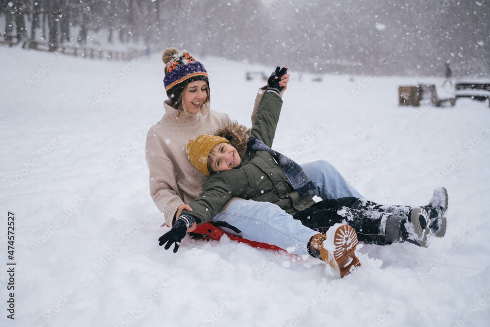 Mother and son having fun in winer snowy park, riding on sled, family travel on vacation, christmas. Winter clothing, winter sports. Lifestyle, kids, parenting, winter nature.