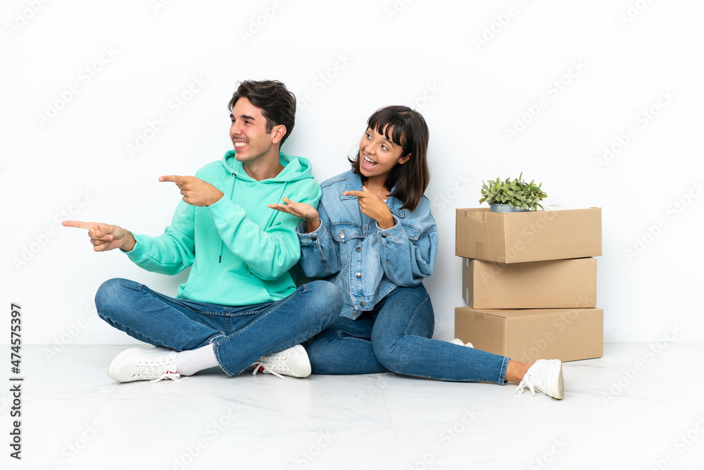 Young couple making a move while picking up a box full of things sitting on the floor isolated on white background extending hands to the side for inviting to come