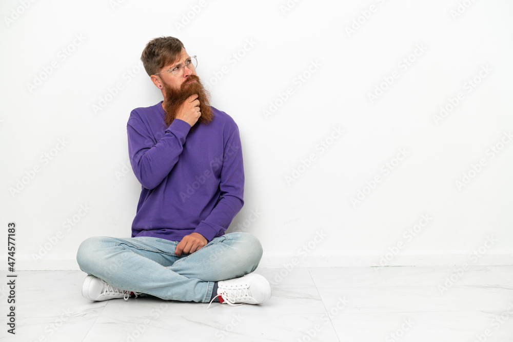 Young caucasian reddish man sitting on the floor isolated on white background having doubts and with confuse face expression Young caucasian reddish man sitting on the floor isolated on white background having doubts and with confuse face expression