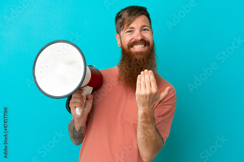 Young reddish caucasian man isolated on blue background holding a megaphone and inviting to come with hand