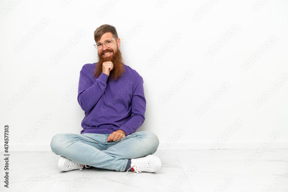 Young caucasian reddish man sitting on the floor isolated on white background with glasses and smiling