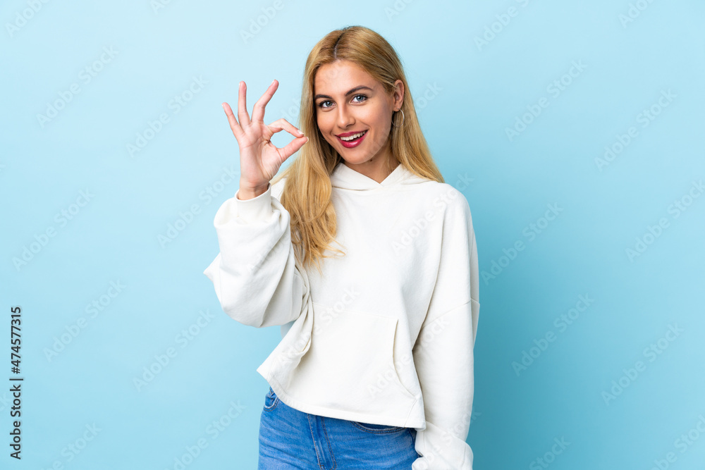 Young Uruguayan blonde woman over isolated blue background showing ok sign with fingers