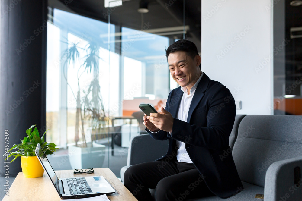 Successful Asian businessman celebrates victory, looks at phone, happy and rejoices sitting in modern office Successful Asian businessman celebrates victory, looks at phone, happy and rejoices sitting in modern office