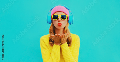 Portrait of stylish young woman in headphones listening to music blowing her lips sends sweet air kiss wearing colorful yellow sweater on blue background