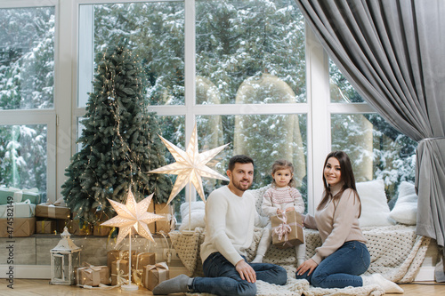 Happy family of mom, dad and little daughter spent time together on the eve of Christmas. Young perents with daughter sits in front of big window. It's snow outside photo