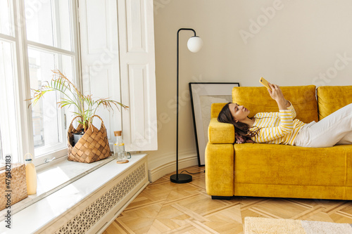 Fotografie Slim caucasian young woman lying on couch using mobile phone app, playing game