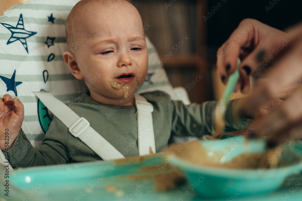 Baby eating with a face stained in food while mother feeds him with a spoon Baby eating with a face stained in food while mother feeds him with a spoon