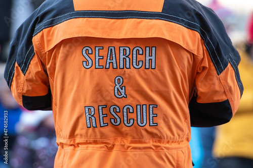 A male adult stands back on wearing a bright orange and black safety jacket that has grey lettering. The safety coat has the words search and rescue in the middle of the industrial uniform.