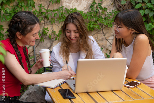 Three friends in the courtyard of a cafe. One writes in her agenda, with a computer and mobiles on the table. Women laughing and talking.