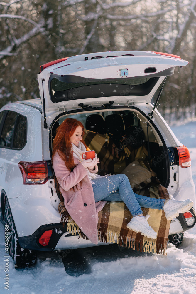 Winter holidays. Joyful young woman drinks hot cocoa while sitting on trunk of car.