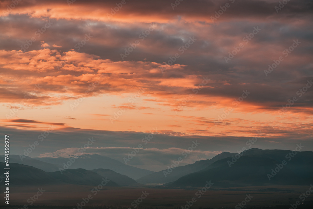 Scenic dawn mountain landscape with orange low clouds in valley among mountains silhouettes under cloudy sky. Vivid sunset or sunrise scenery with low clouds in mountain valley in illuminating color.